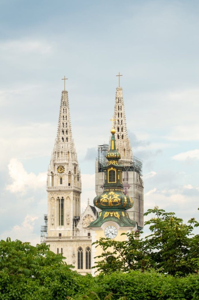 Zagreb Cathedrals gothic towers stand tall against a cloudy sky amidst greenery.